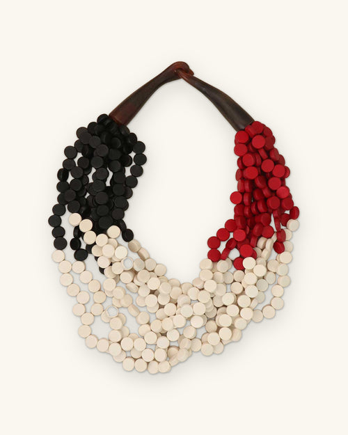 Color-blocked wood disc bib necklace in black, red and cream with a horn closure, made in the Philippines, shown on a sand background